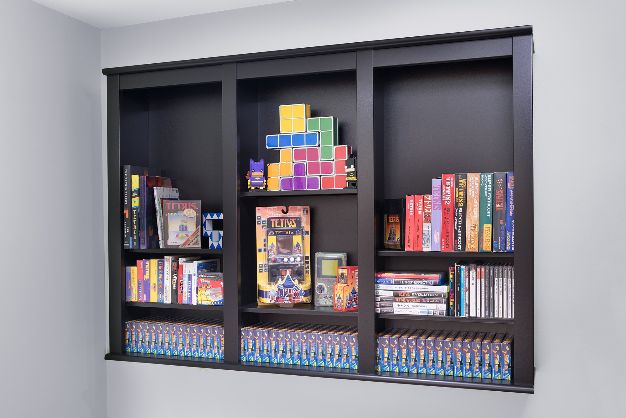A black shelf mounted to a wall with three large vertical sections which are divided into subsections. On the shelves are various Tetris games and Tetris related collectables.